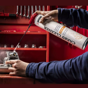 Worker in blue sleeves applies lubricant spray to a small engine component on a red workbench, using a can with a red nozzle.